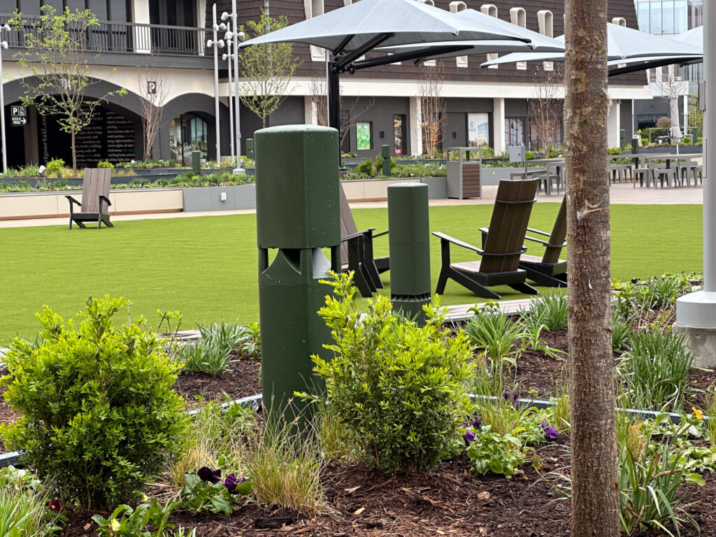 Sound Systems for Outdoor Environments. A landscaped outdoor area with green grass, plants, and shrubs, featuring green cylindrical objects, wooden chairs, and large umbrellas providing shade, set near a modern building.