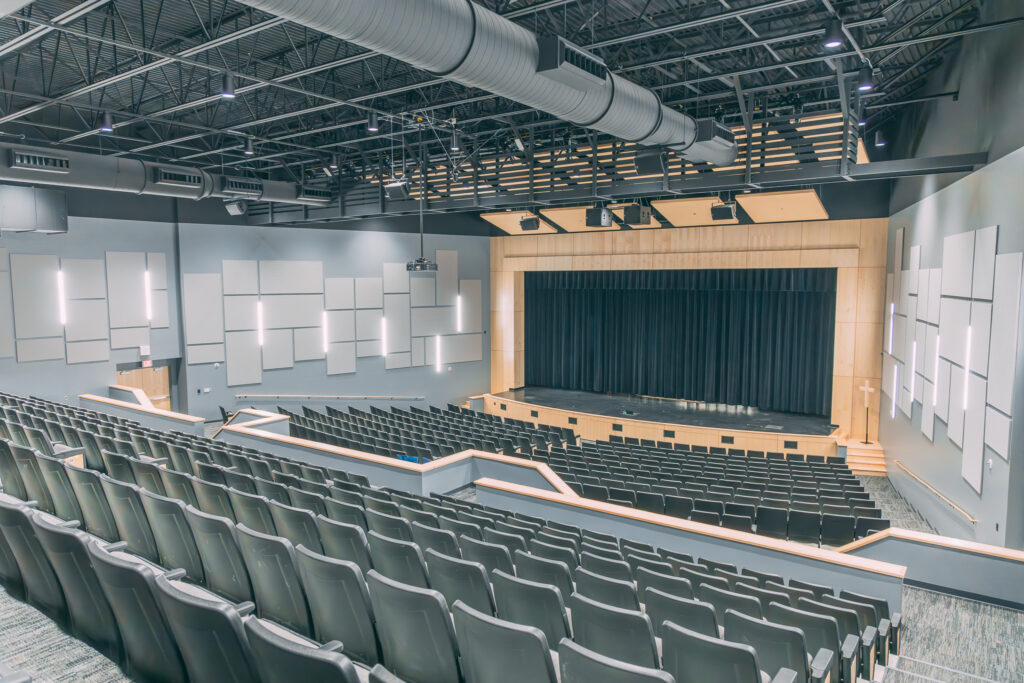 Rows of empty seats in a modern auditorium with a wooden stage and black curtains, surrounded by acoustic panels and overhead lighting, viewed from the rear of the seating area.