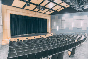 A modern auditorium with rows of empty black seats facing a stage with a closed black curtain. The room features light wood paneling, geometric wall accents, and ceiling-mounted lights and speakers.