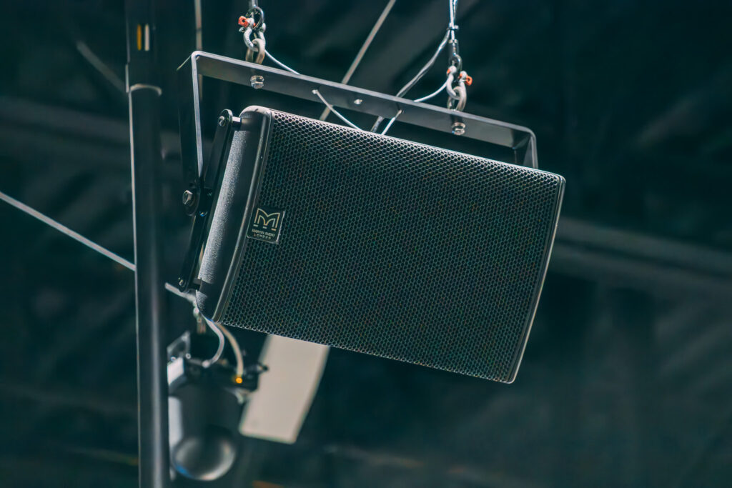 A black Martin Audio speaker is suspended from the ceiling with metal brackets and cables, positioned in an indoor space with an industrial ceiling in the background.