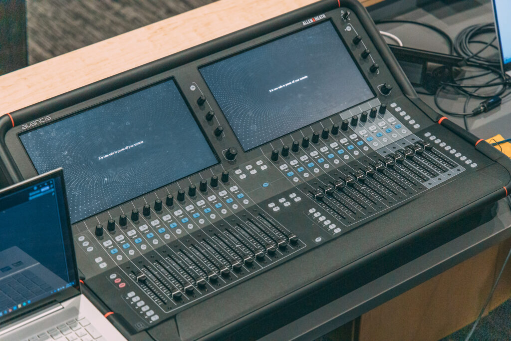 A digital audio mixing console with two large screens, numerous sliders, buttons, and knobs is set up on a desk next to a partially visible laptop. The screens display a simple graphic with minimal text.