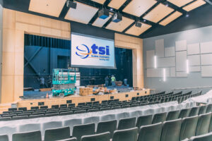 A modern auditorium under construction, with plastic-covered seats, a large projector screen displaying "tsi Global Companies," and workers assembling equipment on stage. Cardboard boxes and tools are scattered around. AV System Installation. Performing Arts Center Construction and Audio System Upgrade, performing arts center