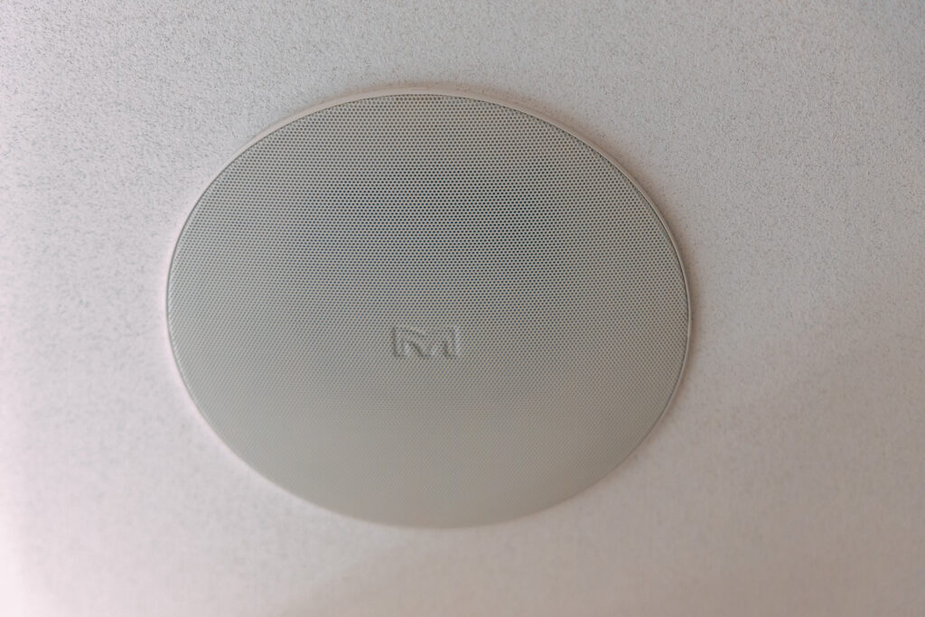 A round, white ceiling speaker with a perforated cover and a small logo in the center, mounted on a textured white ceiling. Intercom System