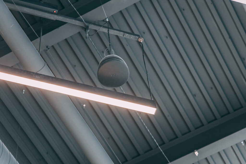 A black speaker and a long LED light fixture are suspended from the ceiling, which has exposed metal beams and a grey industrial design.