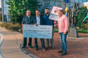 Four men stand outdoors, holding a giant check for $15,000 made out to Shriners Children’s St. Louis from TSI Global. A Shriners Children’s sign, balloons, and a statue are visible in the background. Shriners Childrens Telethon.