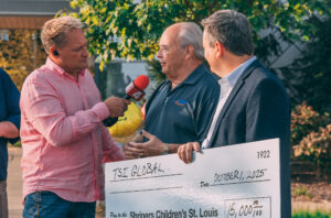 Three men stand outdoors. One holds a microphone and interviews another, who is holding a large ceremonial check for $15,000 made out to Shriners Children’s St. Louis from TSI Global, dated October 1, 2025. TSI Global proudly donated $15,000 at the Shriners Childrens Telethon.