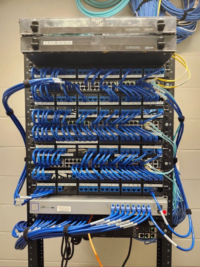 A server rack with organized blue Ethernet cables connected to patch panels and network switches, with Corning equipment on top. Cables run neatly along each row, and the background is a plain wall.