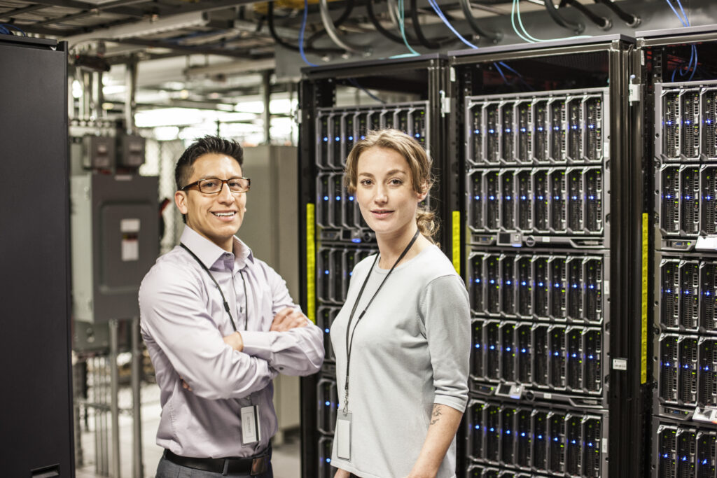 Two IT professionals standing in a server room, smiling at the camera, surrounded by tall racks filled with servers and networking equipment. Data Center Build. 