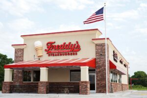 A Freddy’s Frozen Custard & Steakburgers restaurant with a red and beige exterior, a large ice cream cone decoration, and an American flag flying on a pole outside.