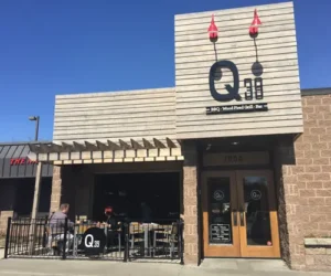 Exterior of Q39 BBQ restaurant with a wooden facade, large sign featuring "Q39," and outdoor seating where two people are dining. The sky is clear and blue.