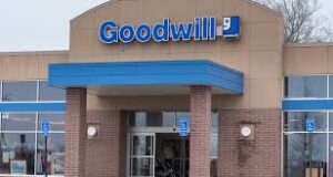The front entrance of a Goodwill store, featuring a large blue sign with the Goodwill logo above glass doors and windows; there are accessible parking signs near the entrance.
