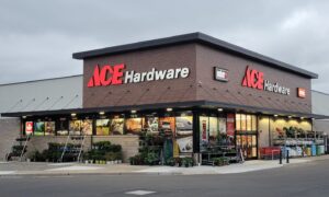 A modern Ace Hardware store with large red and white signage, featuring glass windows displaying products and various plants and outdoor items arranged along the sidewalk on a cloudy day.