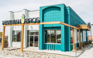 A Hawaiian Bros restaurant with a turquoise and white exterior, large windows, a pineapple logo, and a wooden pergola at the entrance, surrounded by landscaping with rocks and bushes.