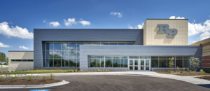 A modern, two-story school building with large glass windows and light gray exterior panels. The letters "RP" are displayed on the upper right corner. The sky is blue with a few clouds and landscaping is in front.